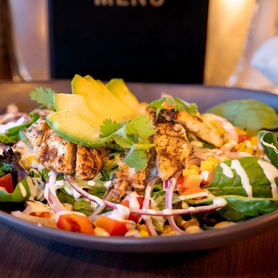 Grilled chicken salad topped with avocado, cilantro, mixed greens, vegetables, and creamy dressing in a dark bowl.