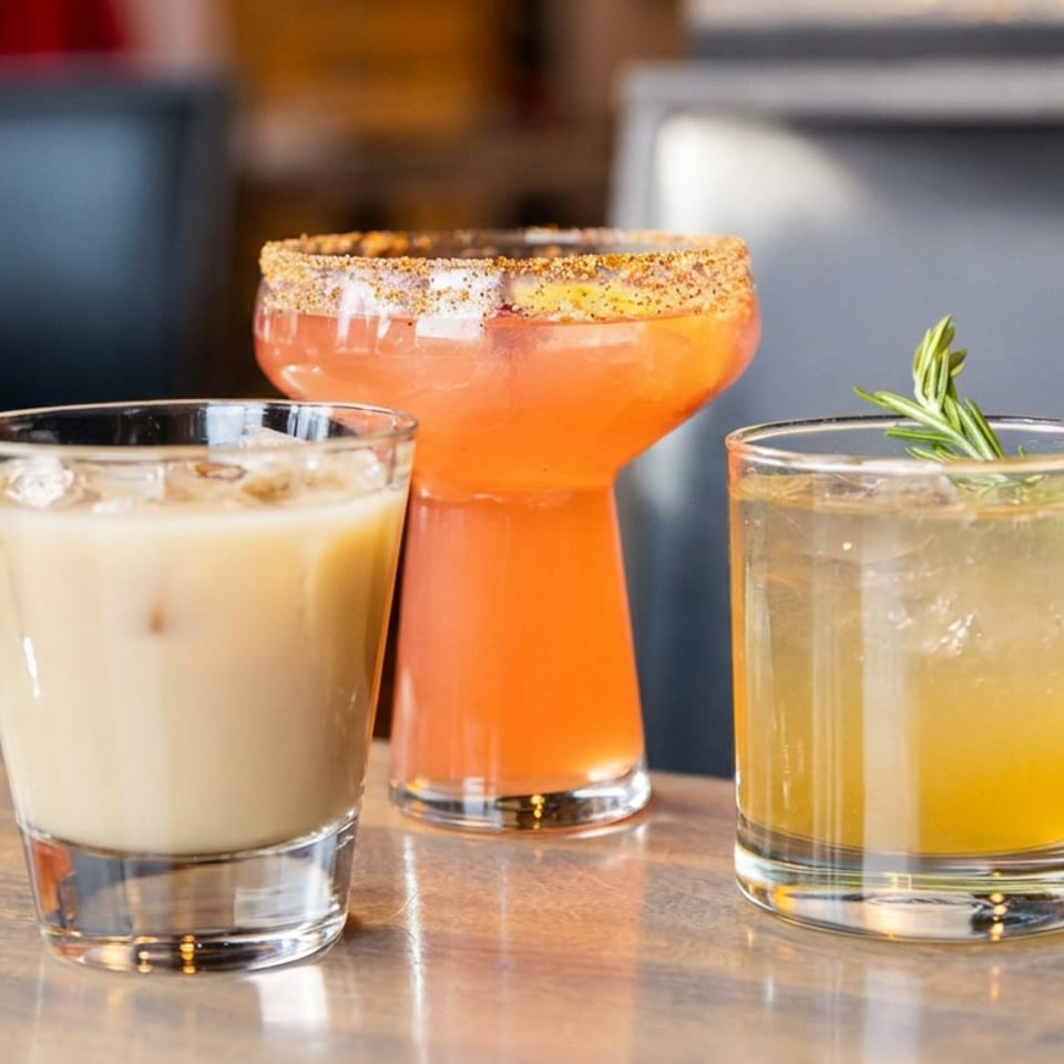Three colorful cocktails sit on a wooden table, including a creamy iced drink, a chili-rimmed orange cocktail, and a rosemary-garnished citrus drink.